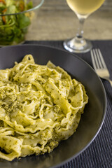Pasta Mafaldine with tofu pesto sauce served on a dark plate. Salad bowl and a glass of wine on textile placemat and wooden kitchen surface.