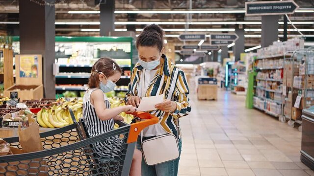 Girl sits in a supermarket trolley and looks at the shopping list. Mother and daughter in the face masks are shopping in a big supermarket. Mothers helper. Shopping during quarantine