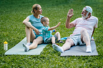 Portrait of happy family. Grandpa, mother and her little son smiling and giving high five sitting on grass outdoor in park meadow. Three different generation concept. - Powered by Adobe