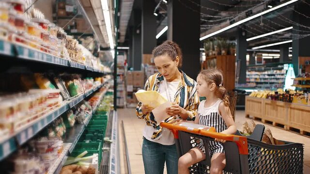 Little girl and her mom, housewife, enjoy family shopping together. A woman takes packed products from the shelf, wrapped mushrooms and puts them in a trolley