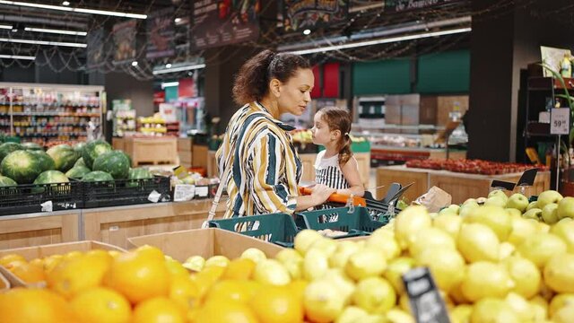 Small daughter sitting in a supermarket trolley. Lovely family, mother and little daughter buy green and red apples in the hypermarket in the fruit section. Vegetarianism and healthy eating concept