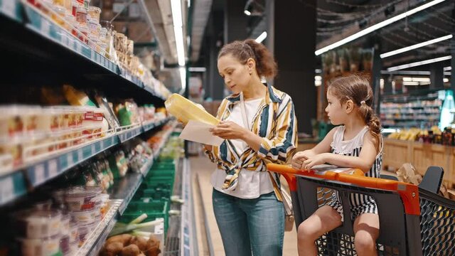 Woman takes packed products from the shelf, packed mushrooms and puts them in a trolley. Little girl and her mom, housewife, enjoy family shopping together