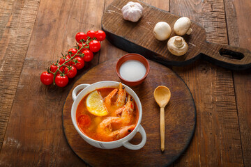 Tom yam soup with shrimps and coconut milk on the table on a round board next to coconut milk mushrooms and tomatoes.