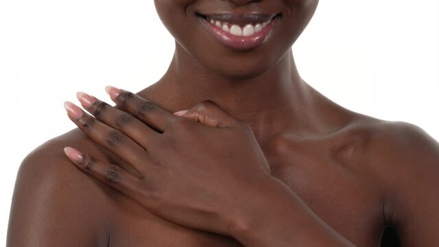 Close up of an African woman's chest and shoulder, facing the camera, applying lotion on her chest and shoulder, smiling. White background.