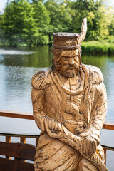 Zwierzyniec, Lubelskie, Poland - 02 July 2021: Wooden sculpture of a traditional dressed man; beautiful pond in the background.