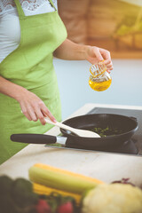 An attractive young dark-haired woman frying meal at worm pan by new keto recipe while standing and smiling in sunny kitchen. Cooking and householding concepts