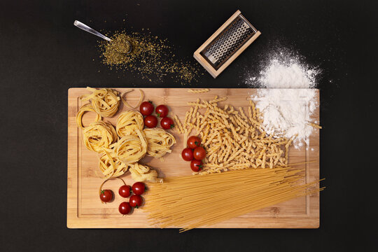 Top view image of a variety of Italian raw pasta, wheat flour, oregano, cherry tomatoes and bamboo table on black background.