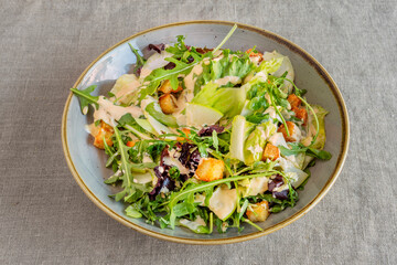 Bowl of green salad with apple slices, cheese slices, croutons, arugula and lettuce sprouts with mayonnaise sauce on gray linen tablecloth