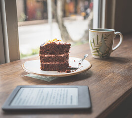 A table with a cup of cooffe a cake, glasses, and an e-reader