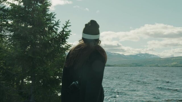Adventurous amateur photographer arriving at a mountain lake while hiking, observing the view. Carrying a hipster camera around her shoulder. Looking thoughtful, seeking beauty. Cinematic movement.