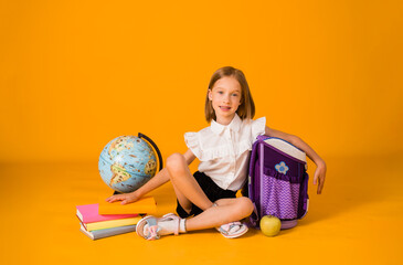 happy schoolgirl in uniforms is sitting with school supplies and a globe on a yellow background...