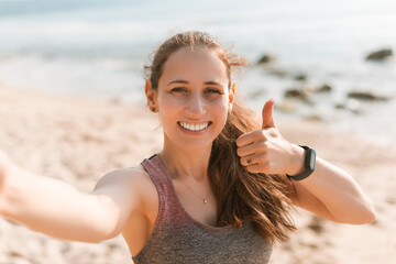 Fototapeta na wymiar Cheerful brunette woman in sportswear making selfie and showing thumb up on the beach at sunrise