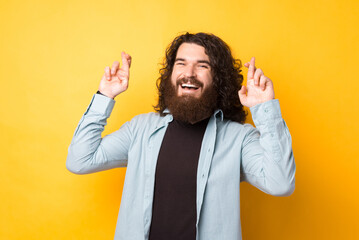 Positive unshaven male with happy expression crosses fingers, hopes for good luck, over yellow background