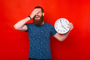 Portrait of bearded younfg man holding clock and keeping facepalm gesture, expressing regret of missed deadline - obrazy, fototapety, plakaty