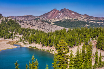 Clear blue alpine lake surrounded by pumice covered moraine, fir trees with a large lava flow and...