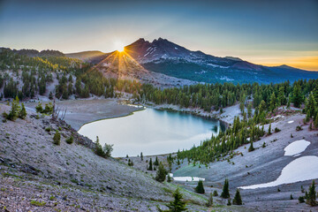 Sunrise over clear blue alpine lake surrounded by fir trees and volcanic mountain peaks.