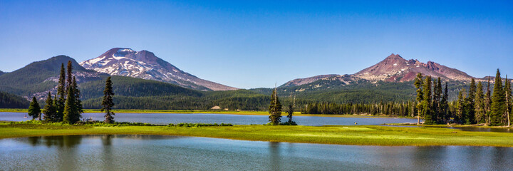 Panoramic view of mountain lake surrounded by fir trees and volcanic mountain peaks.
