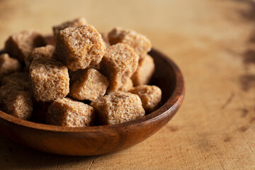 Brown cane sugar cubes in a wooden dish set against a wooden background.