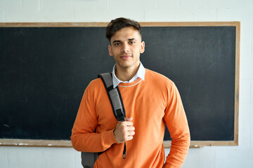 Portrait of young happy confident Indian latin Hispanic high school college university student standing in classroom on black board background, holding backpack on shoulder looking at camera.