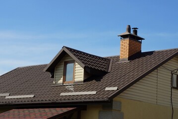 part of a private house from an attic with a window under a brown tiled roof with a brick chimney on the background of a blue house