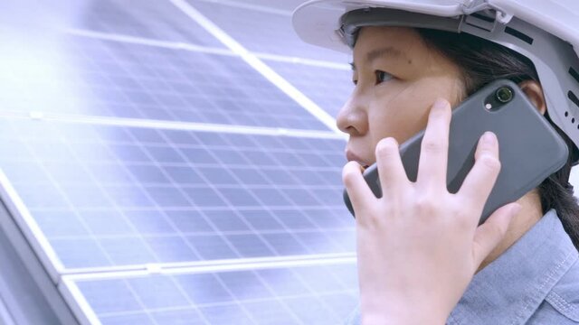 Close up, Asian woman wearing white safety hard hat, working outdoors, making telephone call, talking, nodding beside solar roof. Female engineer or worker and sustainable, renewable energy concept.