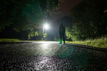 A low angle, shallow depth of field of a spooky hooded figure, silhouetted against street lights. On a rainy country road at night.