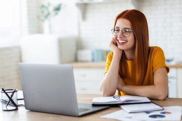 Online education, portrait of red-haired young caucasian female freelancer or woman student sitting on the table with notebook and laptop computer looking at the camera and smiling pleasantly