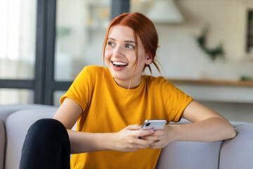 Happy red-haired girl is texting online in social networks, messenger using mobile phone sitting on the couch at home. Online communication, technology concept