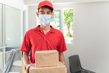 Fototapeta na wymiar Portrait of Asian delivery man in uniform wearing face mask and holding small box package, smiling and looking at camera. Product logistic service and delivery during coronavirus pandemic concept.