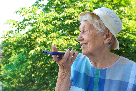 Charming elderly woman in rectangular black rimmed eyewear using voice recognition function on cell phone.
