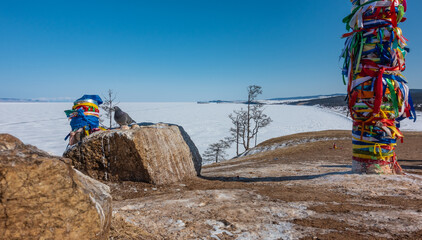 Ritual pillars tied with colorful bright ribbons stand on the hill. Nearby, on a boulder, sits a dove. In the distance you can see the snow-covered frozen Lake Baikal. Clear blue sky. Winter sunny day