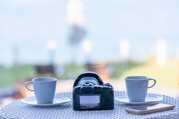 Camera and cups of coffee on a white table in a room with blurred ocean background