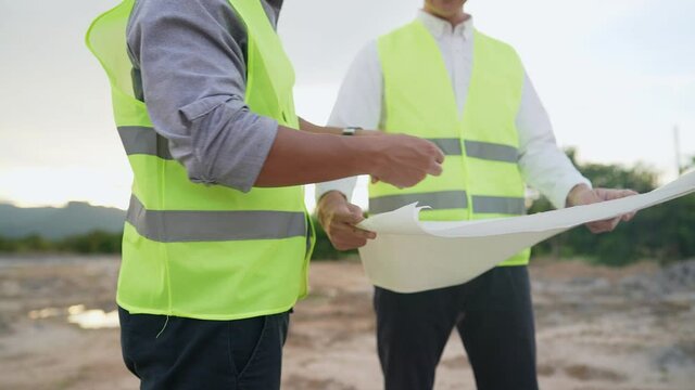 Asian engineer architect employee wear safety vest and safety helmet work together at construction outdoor site, holding tablet blueprints paper, using tape measure, measuring the blueprint perimeter
