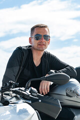 Attractive stylish young man in sunglasses sits on a motorcycle. Portrait handsome biker posing on a bike in a black leather jacket. Life style photography. Vertical shot. Blue sky background