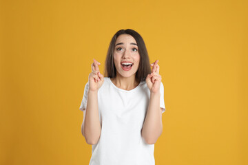 Excited young woman holding fingers crossed on yellow background. Superstition for good luck