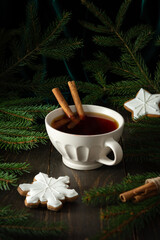 Ceramic mug with black tea and cinnamon among fir branches on a dark wooden background, delicious cookies in the form of a snowflake, Merry Christmas and Happy New Year