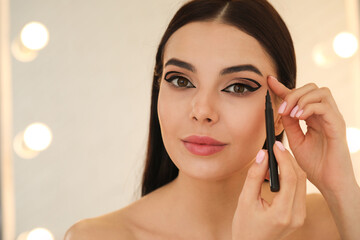 Beautiful young woman applying black eyeliner indoors. Space for text
