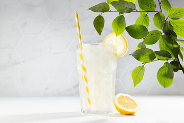 Homemade traditional refreshing cold sour lemonade infused beverage with ice, straw in glass with yellow lemon slice, green leaves in sun beam in modern white kitchen interior