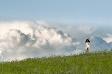 Fototapeta premium asian long haired woman in white dress on the meadow frozen in motion with a raised hand against сumulus clouds