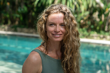 Portrait of young woman with curly long blonde hair and blue eyes and blue swimming pool with garden on background