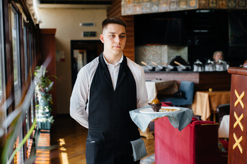 A young waiter in a stylish uniform stands with an exquisite dish on a tray near the table in a beautiful restaurant close-up. Restaurant activity, of the highest level.
