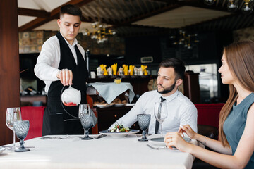 Fototapeta na wymiar A young waiter in a stylish apron serves a table with a beautiful couple in a refined restaurant. Customer service.