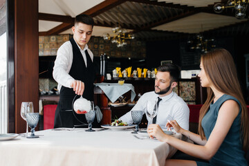 A young waiter in a stylish apron serves a table with a beautiful couple in a refined restaurant. Customer service.