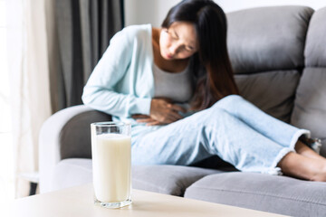 Obraz na płótnie Canvas Unhappy young Asian woman having bad stomach ache with glass of milk on table. Lactose intolerance, food allergy concept.