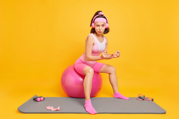 Serious Asian woman being in good physical shape trains hands with resistance band sits on fitness ball listens favorite music via headphones dressed in sportsclothes isolated over yellow background