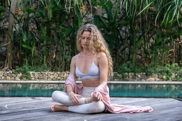 Young woman with curly long blonde hair and blue eyes in beautiful yoga asana next to blue swimming pool and garden