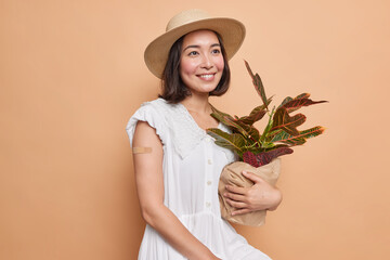 Thoughtful Asian woman with dark hair holds potted plant wears adhesive plaster on arm after...