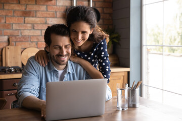 Happy millennial wife giving support to husband working at laptop from home, touching shoulders. Married couple surfing internet on computer together, paying bills online, reviewing mortgage terms