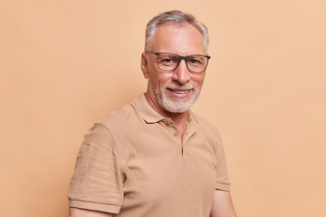 Portrait of handsome bearded senior man smiles pleasantly wears spectacles and casual t shirt looks...