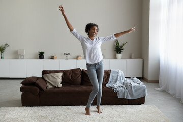 Overjoyed African American woman dancing in modern living room alone, having fun, moving to favorite good music, excited happy young female tenant enjoying leisure time at home, funny activity